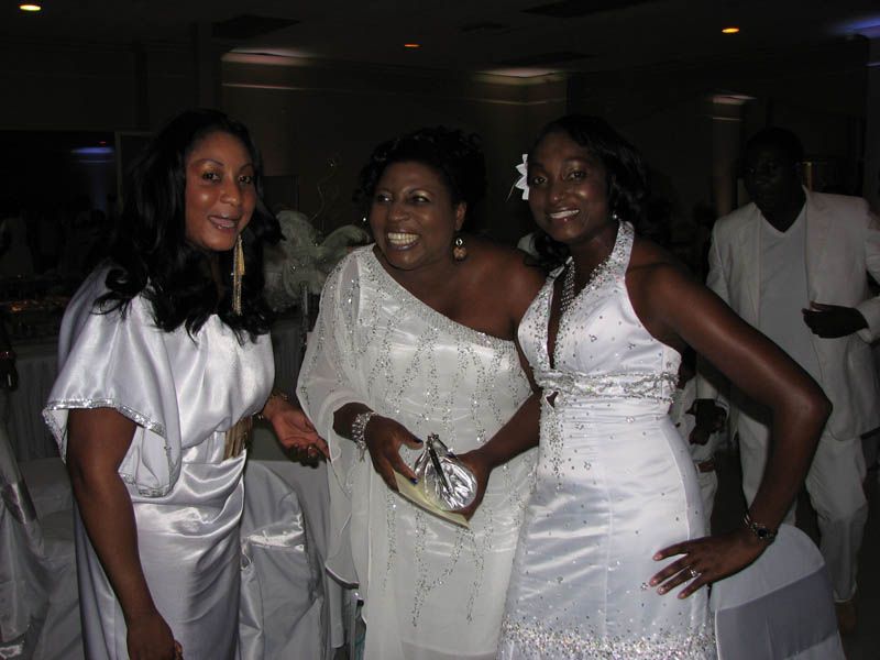 Three women in white formal wear smiling at an event, with one holding a clutch.