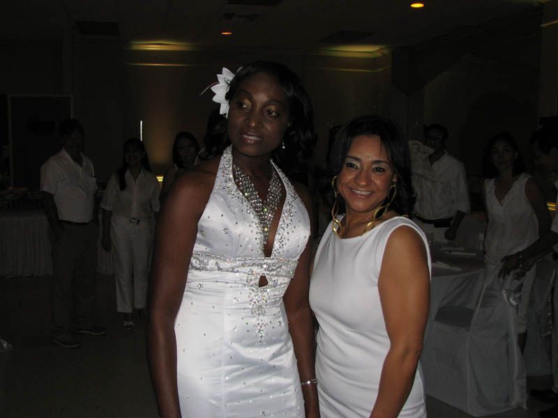 Two women in white dresses smiling in a room with other people.