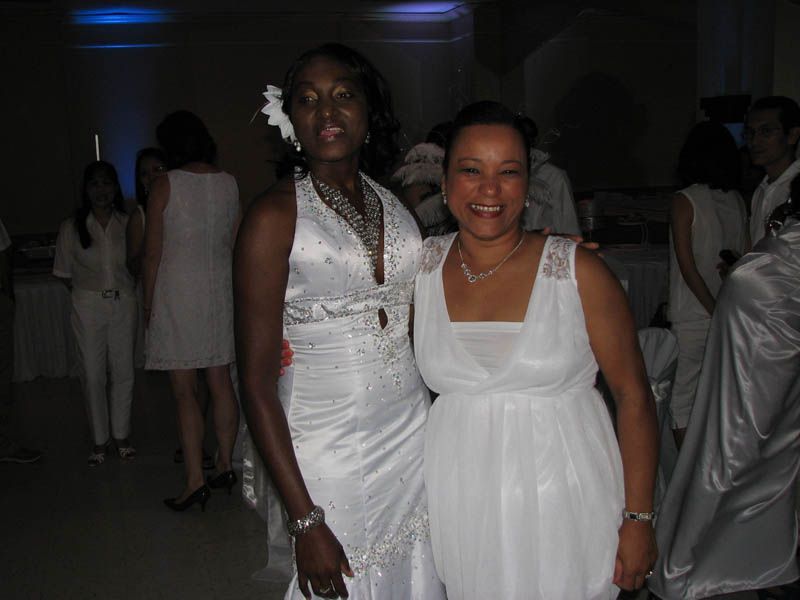 Two women in white dresses pose together at an event. Other guests wear white.