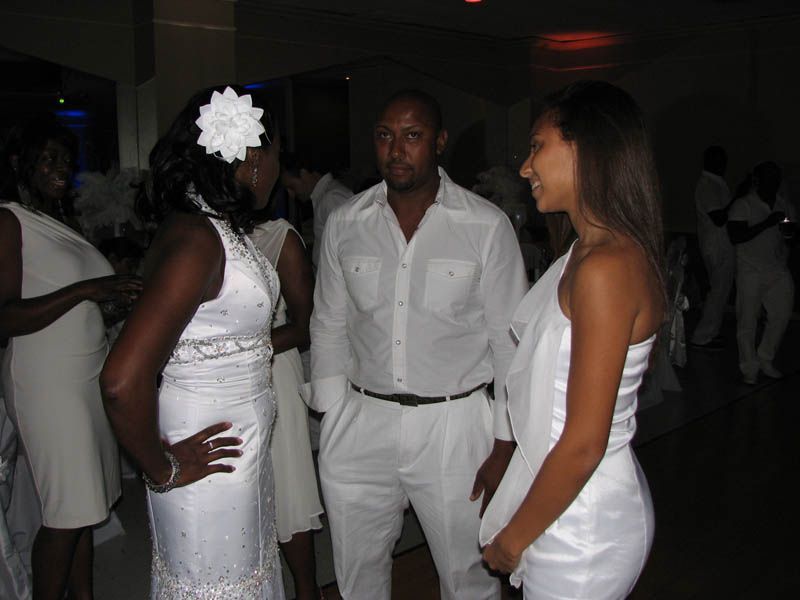 Three people in white attire conversing at a social gathering. Man in the middle; woman on each side.