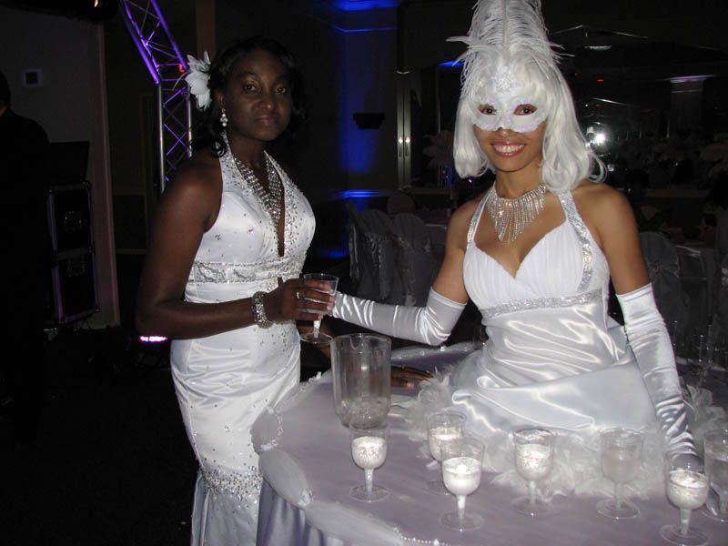 Two women in white dresses at a party. One wears a mask, feathers, and serves drinks.