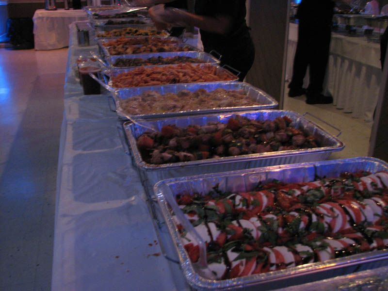 Buffet table with several trays of food, possibly at an event, with people nearby.