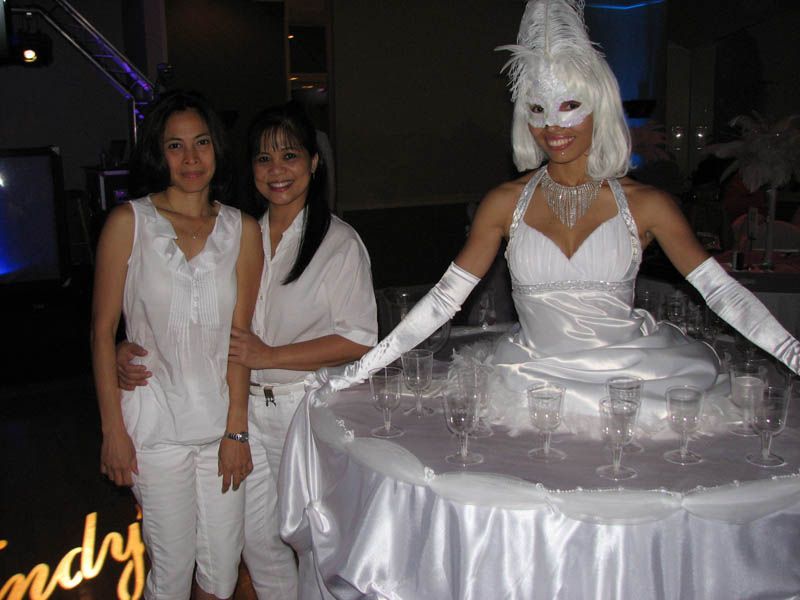 Two women pose with a woman in white costume, carrying champagne glasses on a skirt-like tray.