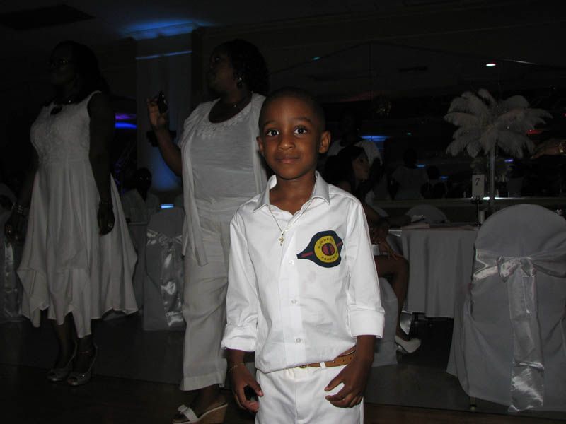 Young boy in white shirt and pants with emblem, standing at a formal event with others in white.