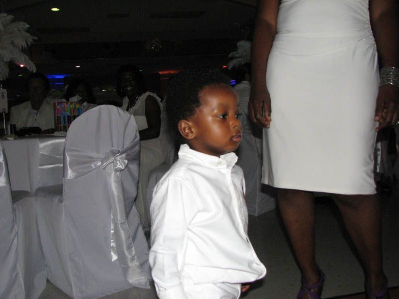 Young child in white shirt stands in a room, looking to the side. A person in a white dress is nearby.