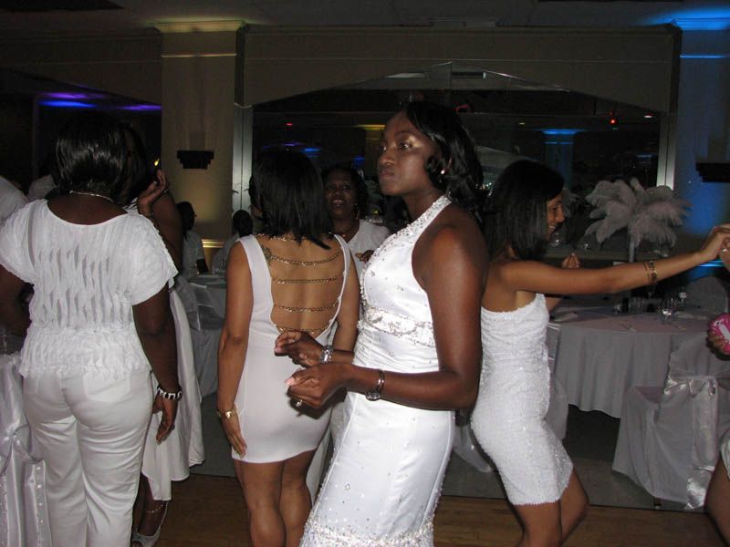 People dancing at a party, mostly wearing white dresses. A venue with tables and a mirrored wall.