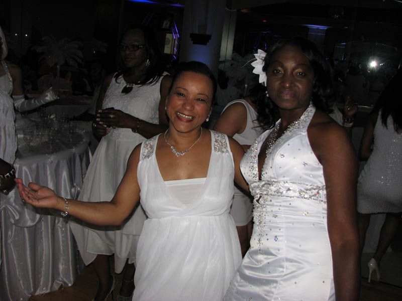 Two women in white dresses smile at a party; others in white are in the background.