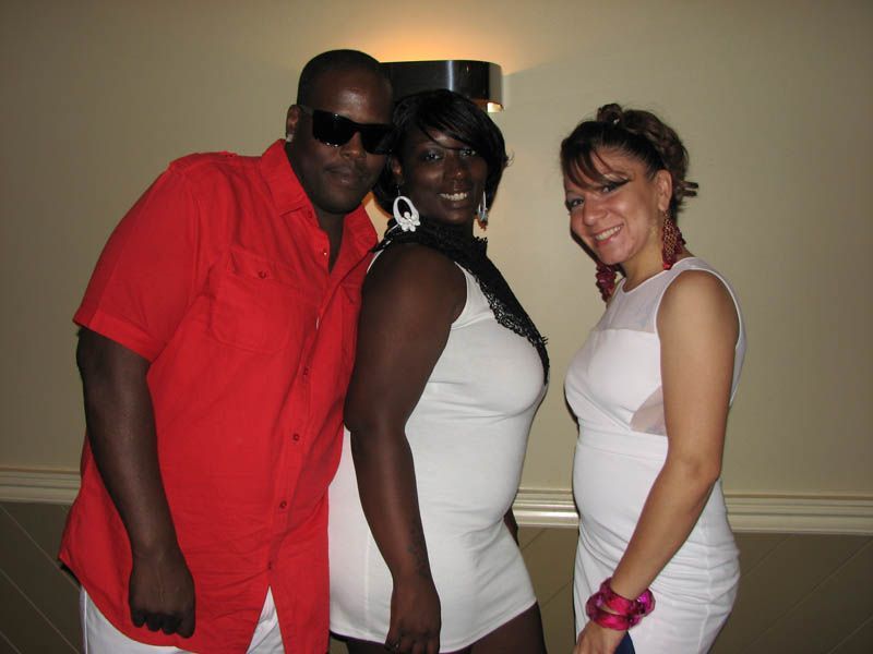 Three people posing. Man in red shirt, two women in white dresses. Indoor setting.