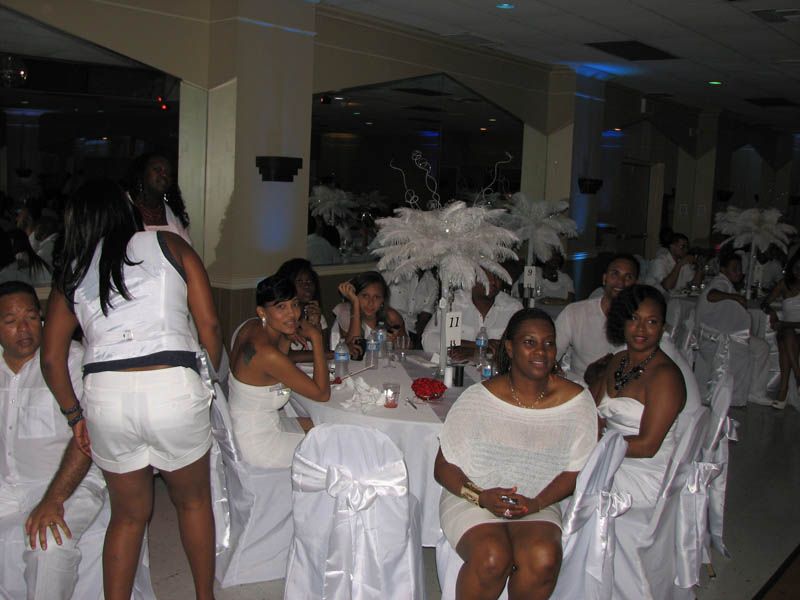 People in white attire at a party seated at round tables. White floral centerpieces adorn tables, in a decorated hall.