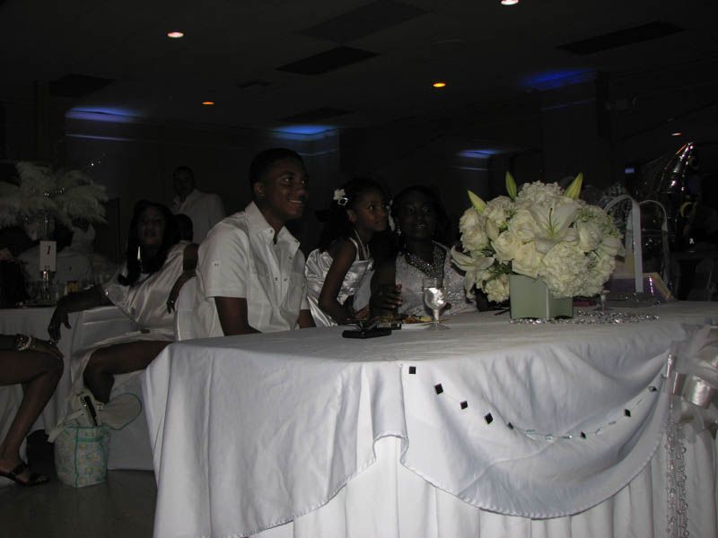 People seated at a table decorated in white, inside a room, a floral arrangement in view.