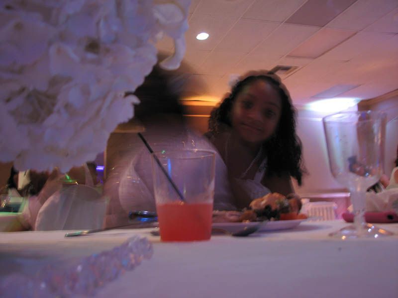 Girl at a table with food and a drink in a blurry setting, party decorations, and pink lighting.