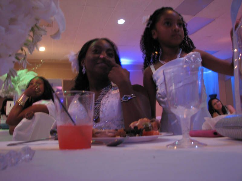 Three people at a table, two looking at camera. One drinks a cocktail. White flowers and blue lighting.