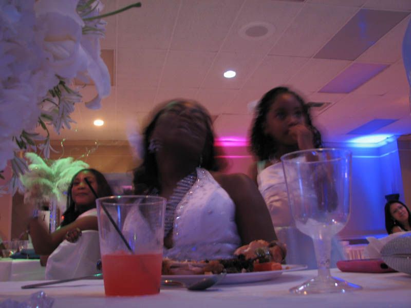 People at a brightly lit event, a woman wearing a white dress is blurry, glasses, food, drinks on a table.