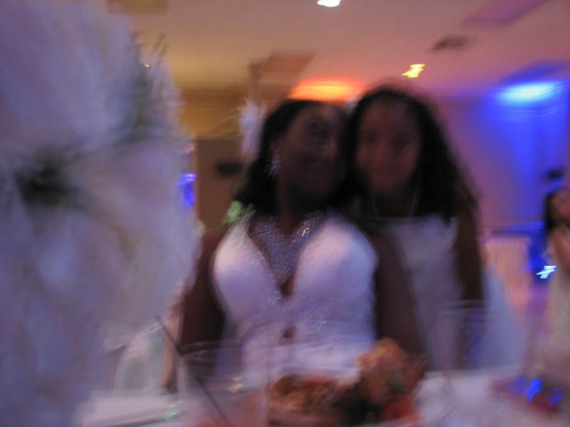 Two people in white dresses at a party with a large white decoration in the foreground, blurred background.