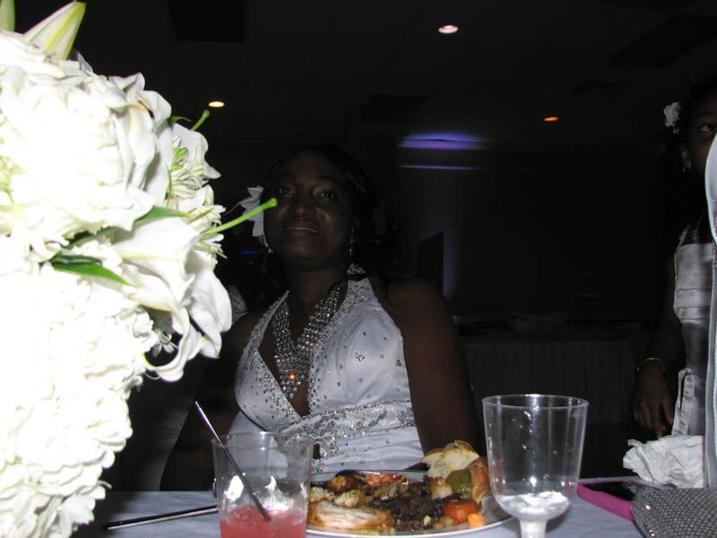 Woman at table wearing white dress, smiling, with food and flowers at an event.