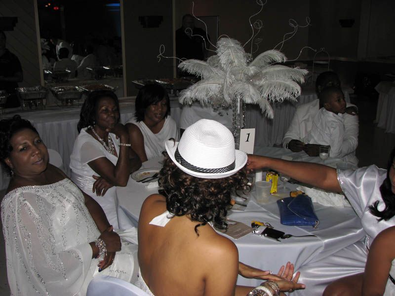 People in white attire at a table with centerpiece. Someone's back is to the camera, wearing a white hat.