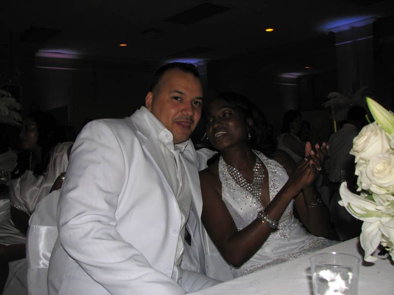 Man and woman in white outfits at a celebration, smiling, with a flower arrangement and table setting in a dimly lit room.