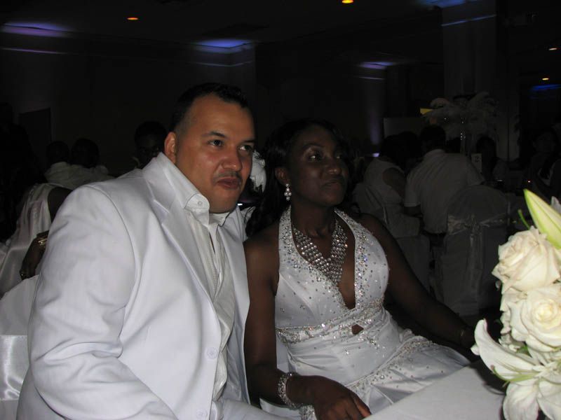 Couple in white formal attire at event. Man in a suit and woman in a dress, with jewelry.