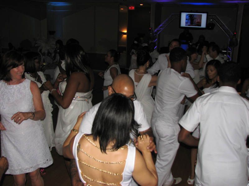 People dancing at a party, all wearing white clothing. Dimly lit room.