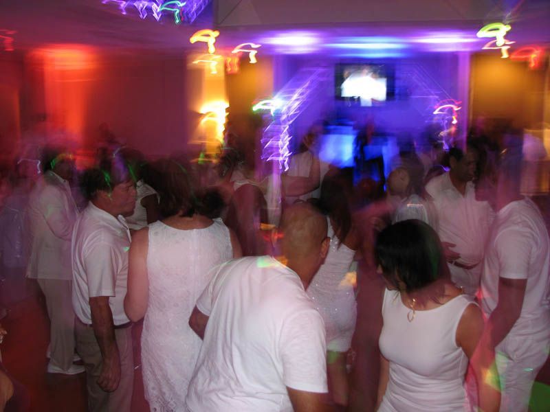 People dancing in a brightly lit nightclub. Most wear white clothes; colorful lights illuminate the scene.