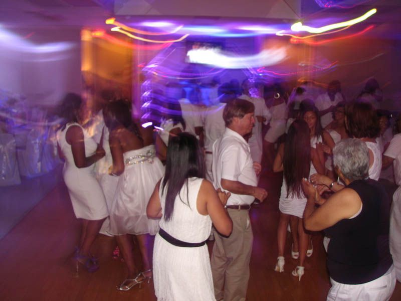 People dancing at a party, mostly wearing white clothes, indoors with colorful lights.