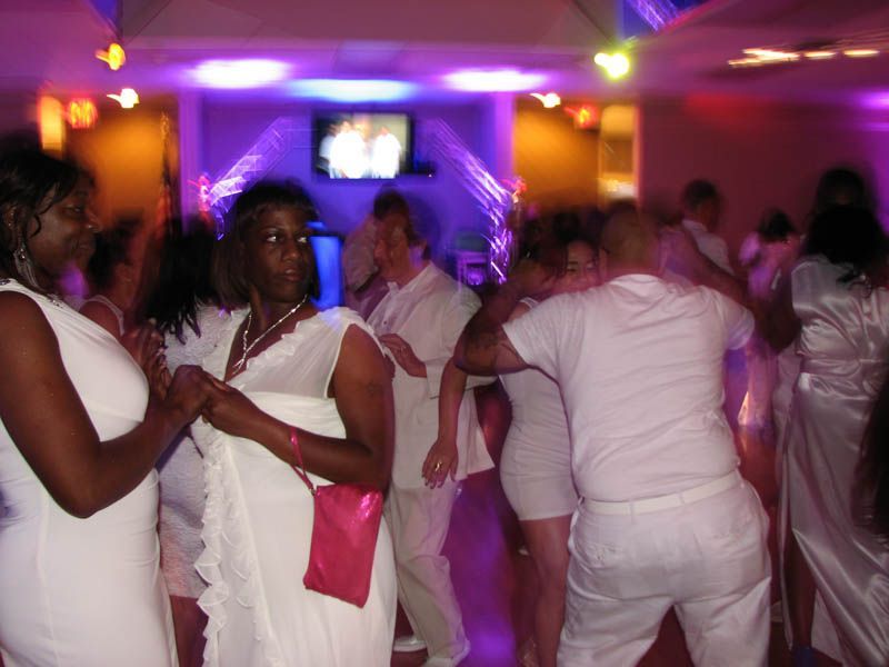 People dancing at a party in a room lit with purple and blue lights; most wearing white clothing.