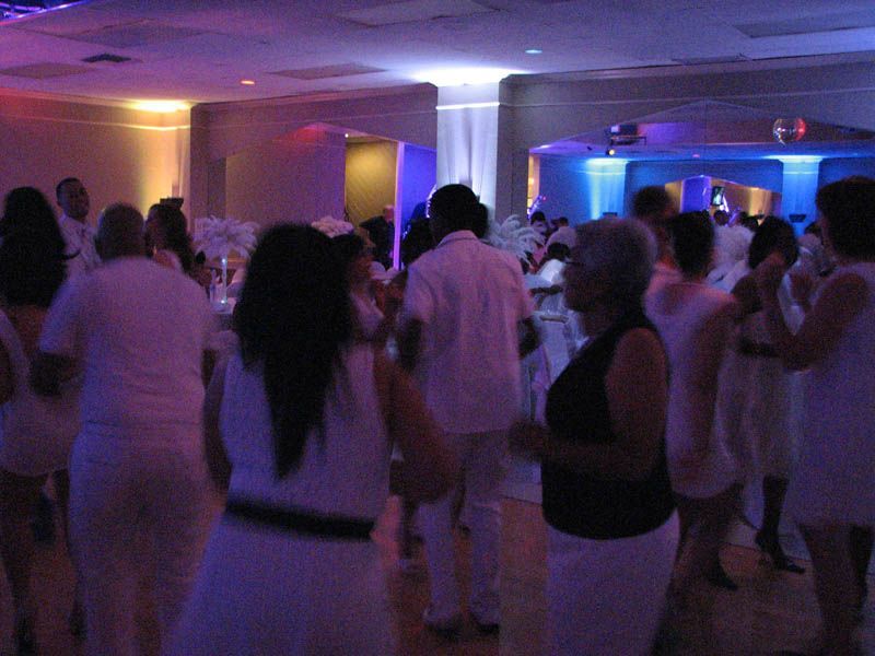 People dancing in a brightly lit room; most are wearing white. Purple and orange lights illuminate the space.