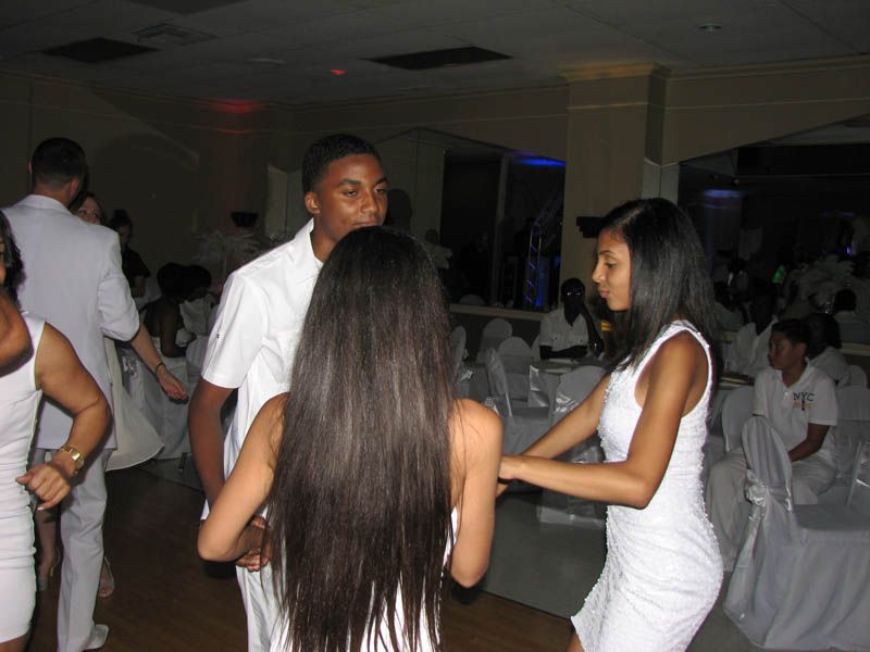 People in white outfits dancing in a ballroom, some with their arms around each other.
