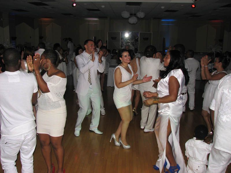 People in white outfits dancing at a party in a large hall with wooden floor.