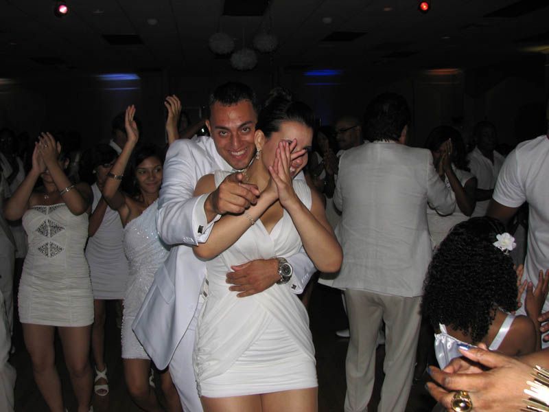 People in white outfits dancing in a brightly lit room. A man embraces a woman, both smiling. Other dancers applaud.