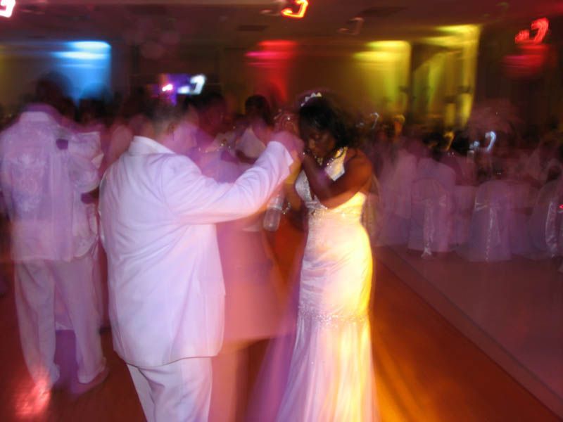 Couple dancing at a lit wedding reception. The bride in white gown, groom in white suit, other guests in white.