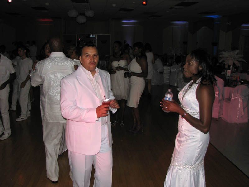 A group of people in white attire at an event, a man in a pink suit and a woman in a white dress are in the foreground.