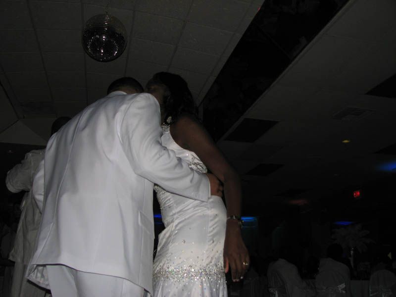 Couple dancing close at an event. Man in white suit, woman in white dress, disco ball overhead.