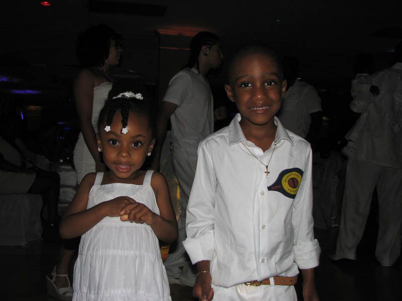 Two children in white outfits smile at the camera at an event with other people.