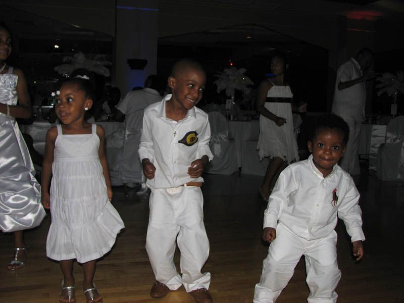 Children dancing at an event, wearing white outfits. They are indoors in a large hall with other people.