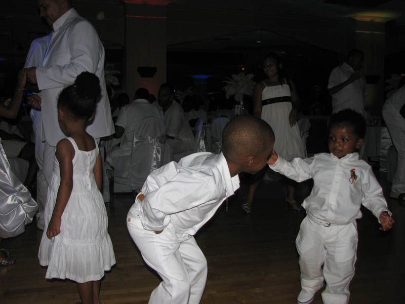 Children in white outfits interacting on a dance floor. Other people in white attire visible in background.