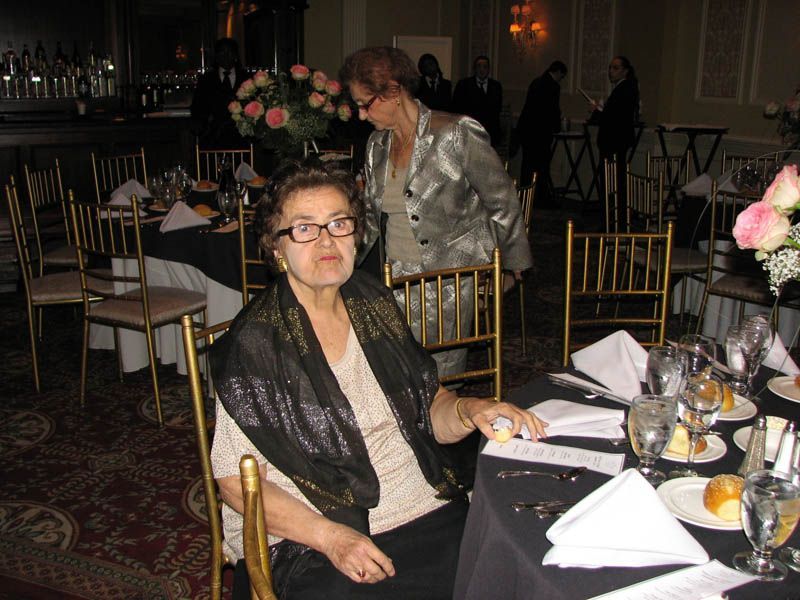 Woman seated at a table in a formal setting, wearing glasses and shawl; another woman stands behind her.