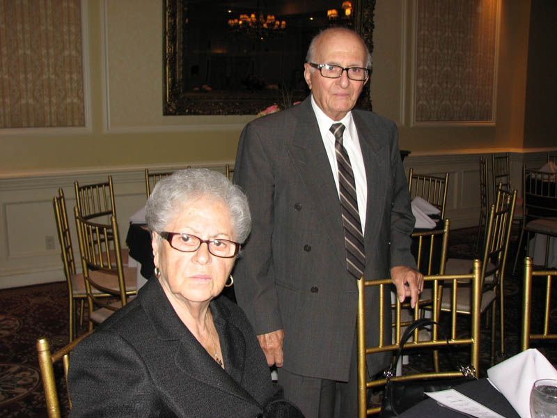 Couple in formal attire at a table in a banquet hall. The woman is seated, the man standing.