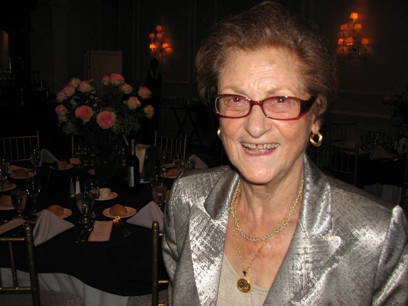 Woman with glasses smiles at event, wearing silver jacket and gold jewelry, floral centerpiece visible.
