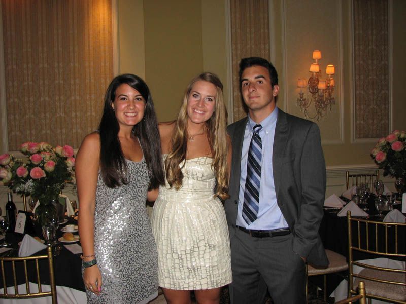 Three people pose at an event: a woman in a silver dress, a woman in a cream dress, and a man in a suit.