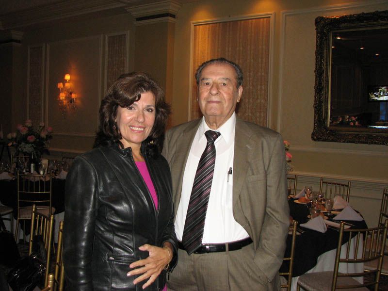 Woman in leather jacket and man in suit stand in a room with tables and a large mirror.