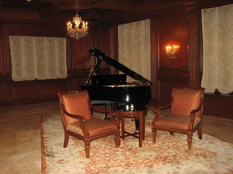 Grand piano in a wood-paneled room, with two armchairs and a small table on a patterned rug.