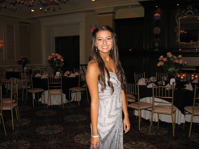 Woman in a silver beaded gown smiles in a ballroom with round tables set for a formal event.