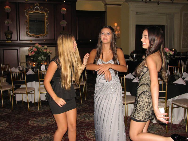 Three young people in formal attire dancing at a party with tables and decorations.