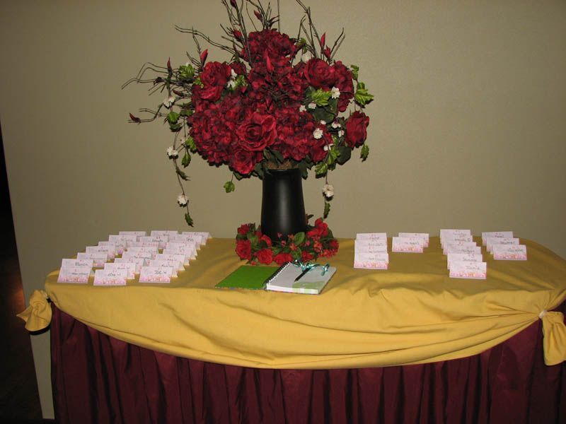 Table with flower arrangement, place cards, and guest book. Gold tablecloth, red skirt.