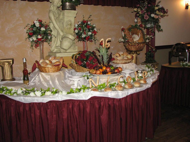 Buffet table with fruit, bread, and floral arrangements. Burgundy tablecloth, gold accents, and a statue.