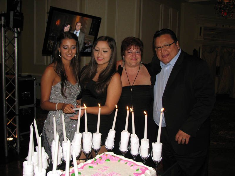 Family celebrates birthday with cake and candles. Black dresses, smiles. Dimly lit room.
