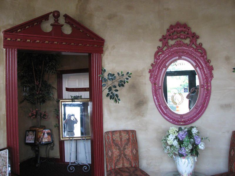 Red-framed doorway and mirror reflect person; wall has floral decor and another decorative mirror, chairs.