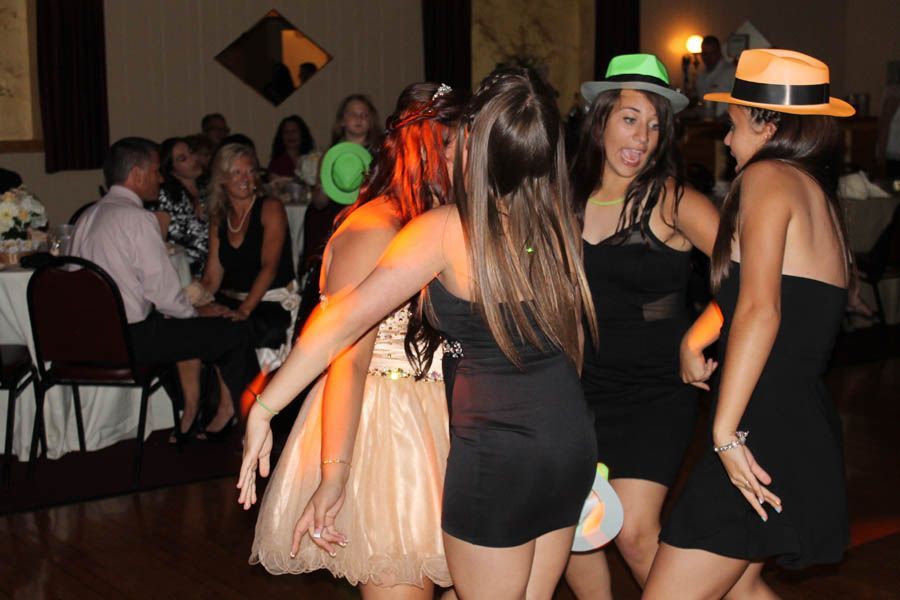 Four young women dance at a party, wearing hats and short dresses. Guests sit at tables in the background.