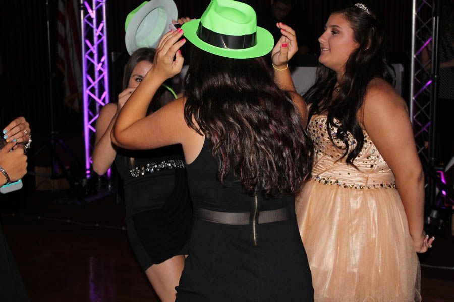 Three young people dance at a formal event, wearing hats and dresses. Purple stage lights illuminate the dance floor.